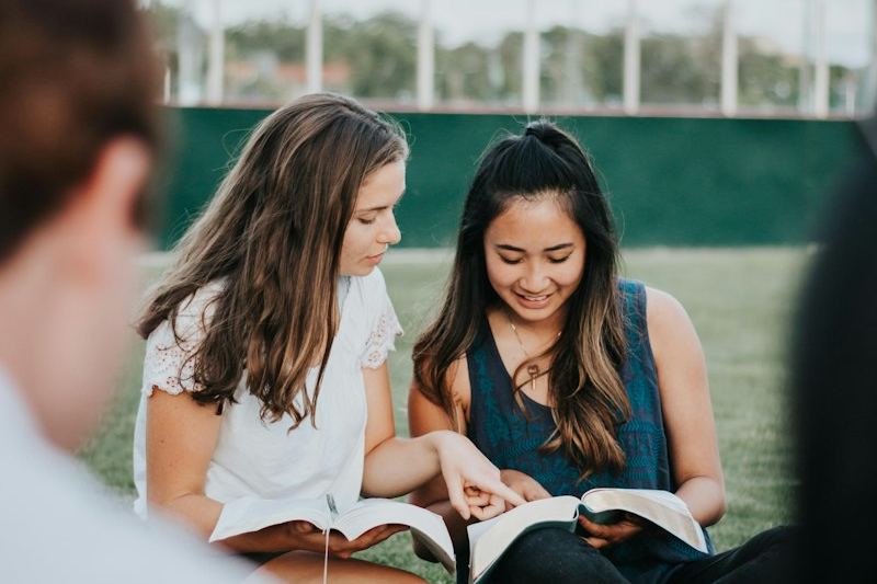 Two young women sitting on the grass with open Bibles, studying together