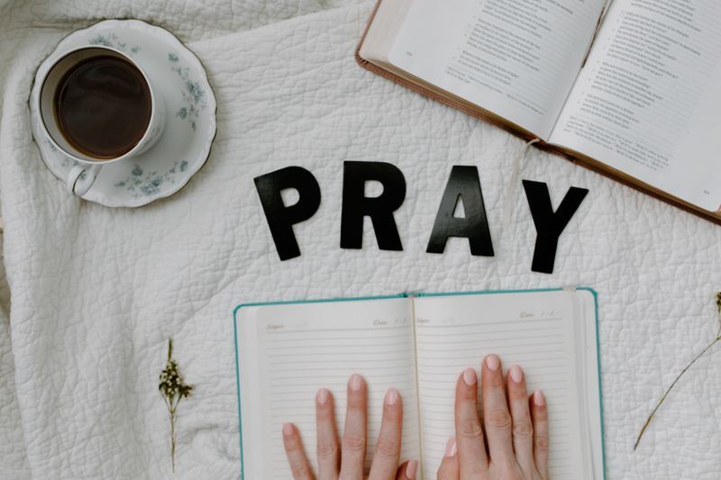 Hands resting on an open journal beside a Bible, a cup of coffee and the word PRAY